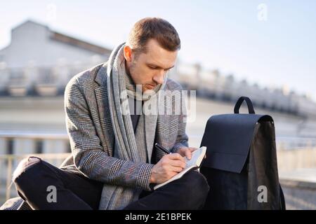 Caucasian bearded man in scarf and grey coat making notes outdoor. Male thinking and working on a sunny spring or autumn day. Working and studying con Stock Photo