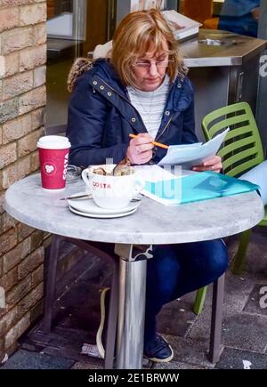 A lady sits at an outside table at Costa Coffee, with a look of concentration & pen & paper in her hand. Papers & folder on table. Shoreditch, London Stock Photo