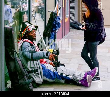 Man in pirate hat sits on pavement in front of shop, next to personal possessions, talking to young woman. Shoreditch, London. Stock Photo