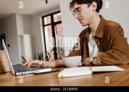 Picture of positive handsome man indoors at home using laptop computer and having a breakfast Stock Photo