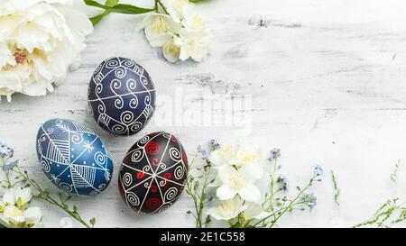 Three handmade painted Easter eggs with spring flowers. Ukrainian pysanka decorated with wax-resist dyeing technique on white shabby wooden background Stock Photo