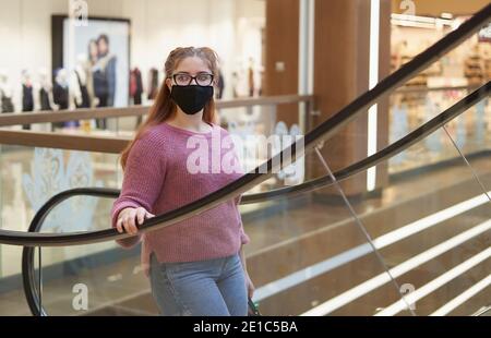 Young woman in a protective mask climbs the escalator in the mall Stock Photo