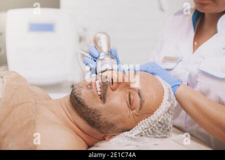 Relaxed handsome mature man smiling, getting ultrasound cavitation treatment by professional cosmetologist. Beautician using hardware cosmetology equi Stock Photo