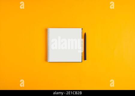 Blank spiral notepad or notebook in square shape with black pen next to it on yellow background. Stock Photo