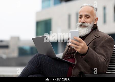 Senior businessman with laptop using smartphone outdoors Stock Photo