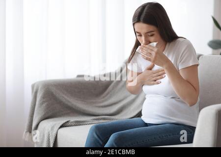 Sick pregnant woman sitting on couch and coughing into tissue Stock Photo