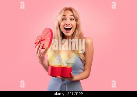 Celebrating Valentine's Day. Happy blonde woman with open heart shaped gift box looking at camera on pink background Stock Photo