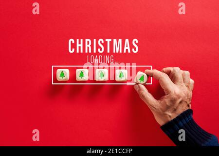 Male hand putting wooden cubes with tree icons in Christmas loading bar on red background. Christmas countdown or celebration concept. Stock Photo