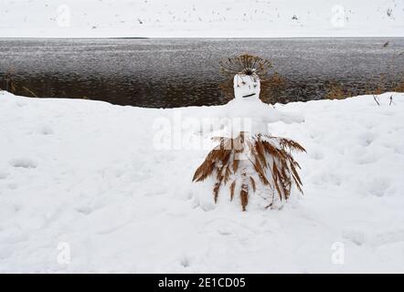 A sad snowman in a cane skirt stands on the river bank on a winter day. Stock Photo