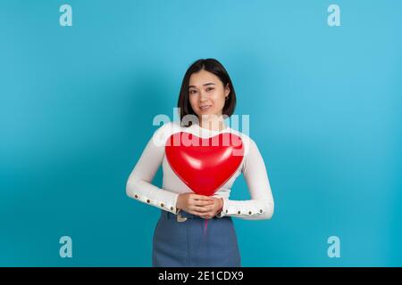 close up a beautiful young smiling Asian woman holds a red heart-shaped balloon near her chest, isolated on a blue background Stock Photo