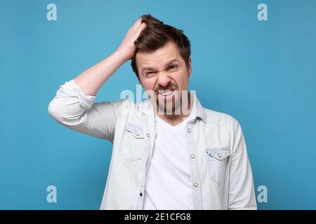 Confused young bearded manscrathing his head being puzzled. Studio shot on blue wall. Stock Photo