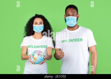 Volunteers Wearing Masks Holding Globe Posing Gesturing Thumbs-Up, Green Background Stock Photo