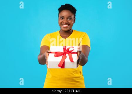 Cheerful Plus-Size African Woman Offering Gift Box Posing, Blue Background Stock Photo