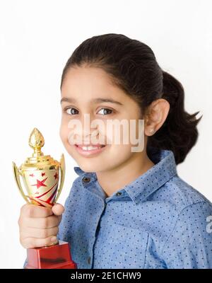 Portrait of a beautiful and smiling Indian school girl in white background holding a golden trophy and depicting victory. Stock Photo