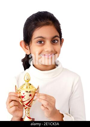 Portrait of a little indian girl in white background with golden winners trophy in hand depicting victory symbol Stock Photo