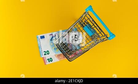 Bitcoin inside small shopping trolley and euro banknotes on yellow background,overhead shot Stock Photo
