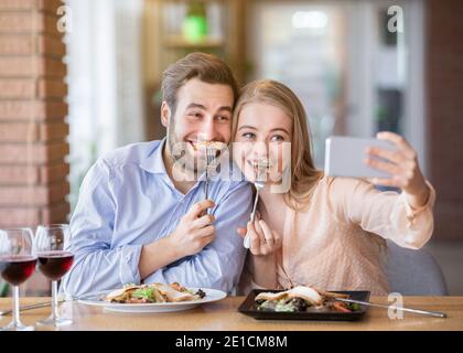 Silly millennial couple having fun during dinner, taking selfie at cozy urban cafe Stock Photo
