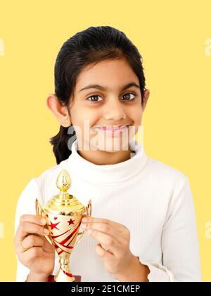 Portrait of a beautiful and smiling Indian school girl in yellow background holding a golden trophy and depicting victory. Stock Photo