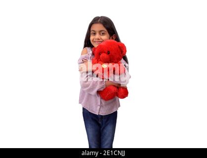 Portrait of a cute little Indian girl hugging her favourite red teddy bear and smiling in white studio background. Stock Photo