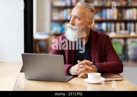 Pensive grey-haired businessman using laptop at cafe, looking through window Stock Photo
