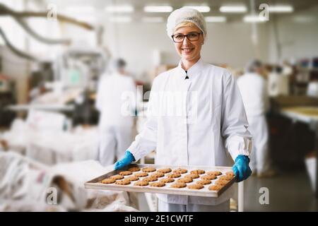 Young satisfied female worker in sterile clothes holding fresh made cookies in food factory. Stock Photo