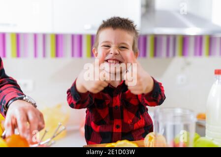 Cute little boy shoving thumbs up. Sitting at kitchen table full of fresh fruits and smiling. Stock Photo