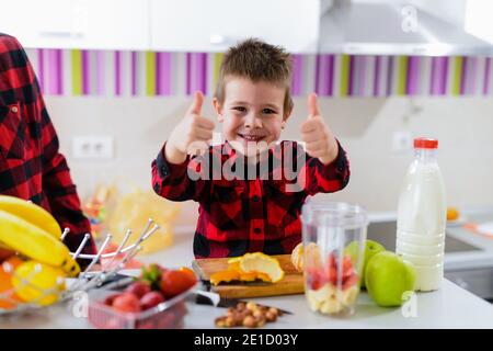 Cute little boy shoving thumbs up. Sitting at kitchen table full of fresh fruits and smiling. Stock Photo