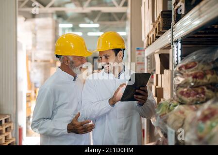 Two coworkers talking and smiling while standing in warehouse. Younger one using tablet. On heads helmets. Stock Photo