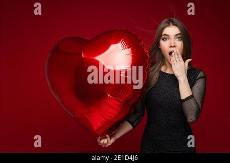 Pretty open-eyed lady with red balloon covering her mouth by hand Stock Photo