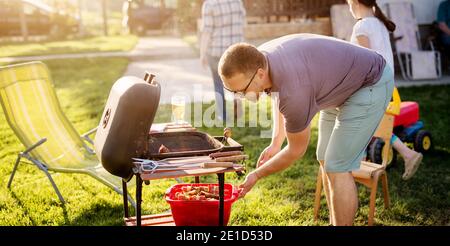 Mature man is taking care of the grill while his family is enjoying in backyard. Stock Photo