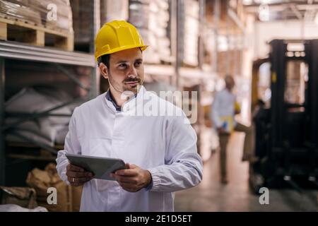 Picture of young focused male manager standing in factory and holding tablet. Dressed in white coat with helmet on his head. Looking satisfied and pro Stock Photo