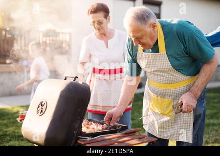 Elder happy couple are checking meat on a barbeque while their family is enjoying in a backyard Stock Photo
