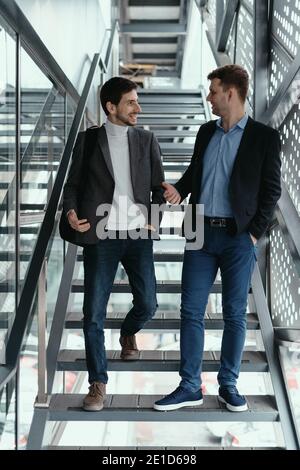 Two young businessmen going up, down the stairs while chatting in a modern building. Stock Photo