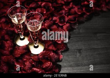 Two drinking glasses of sparkling wine for romantic toast on red rose's petals and wooden table as copy space. Stock Photo