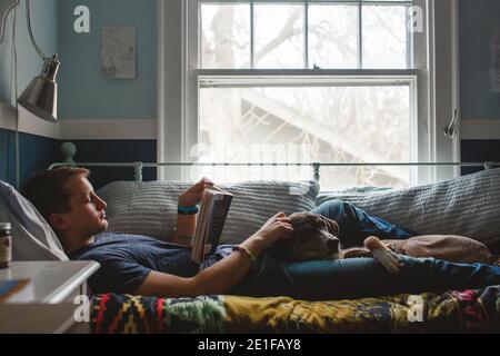 A young man lays on his bed with dog on lap reading by window light Stock Photo