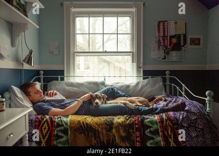 A smiling young man holding book and laying on bed rubs dogs ear Stock Photo