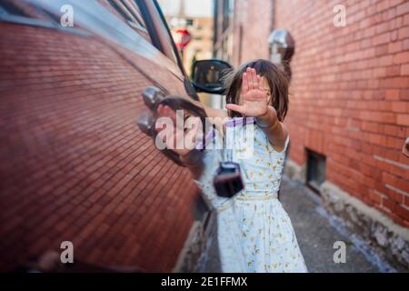 A little girl reflected in side of car holds hand up in front of face Stock Photo