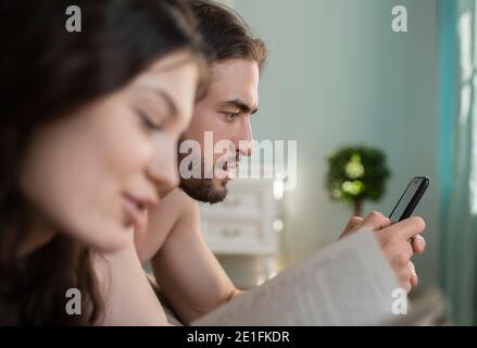 Bearded male using smartphone while resting near reading female in morning at home