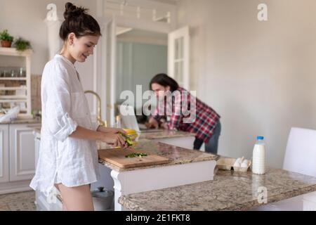 Young female cutting fresh cucumber while preparing salad near working man at home Stock Photo
