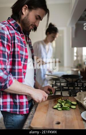 Young male cutting fresh cucumber while preparing food with woman at home together Stock Photo