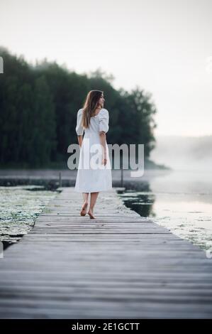 Back view of young woman in white dress standing alone on footbridge and staring at lake. Foggy chilly morning with a mist over water. Stock Photo