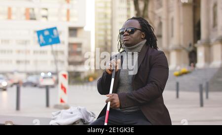 Blind black man sitting on the bench and holding walking long cane. High quality photo Stock Photo