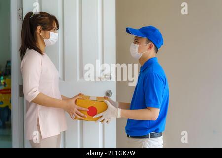 Asian delivery man wearing face mask and glove in blue uniform holding a cardboard boxes in front house and woman accepting a delivery of boxes from d Stock Photo