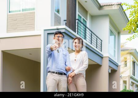 Portrait of Asian young couple standing and hugging together and holding house key looking happy in front of their new house to start new life. Family Stock Photo