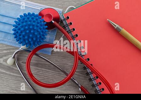 Virus pandemic and medical themed still life with a colorful red stethoscope and surgical face mask placed to the side of wire bound notepad and pen v Stock Photo