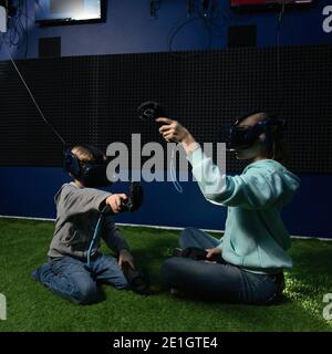 two children wearing virtual reality headset and with joysticks play video games sitting in play area Stock Photo
