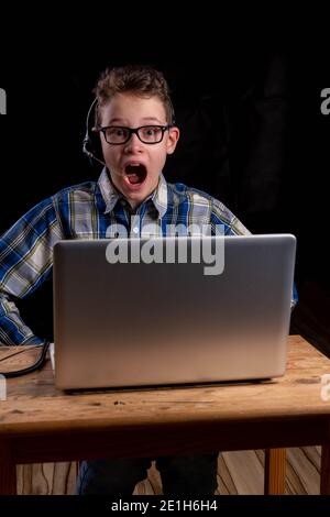 Boy with shirt and glasses joyfully with open mouth and headset doing e-learning on laptop Stock Photo
