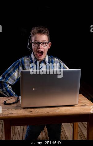 Boy with shirt and glasses joyfully with open mouth and headset doing e-learning on laptop Stock Photo