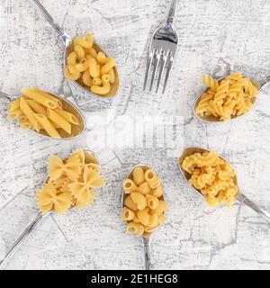 Square image with different types of Farfalle, Pasta a Riso, Orecchiette Pugliesi, and Farfalle pasta in spoons and empty fork on a flat white stone b Stock Photo