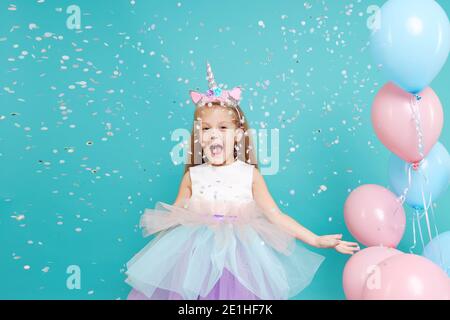 Unicorn girl. Joyful child girl in elegant tulle dress and unicorn headband celebrates birthday  party and blowing multicolored confetti on cyan backg Stock Photo